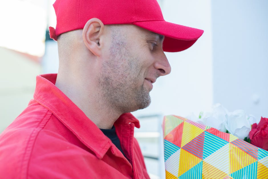 A delivery person wearing a red cap and red jacket is holding a colorful gift box with a geometric pattern, filled with fresh white and pink flowers, during a flower delivery at an outdoor setting wit