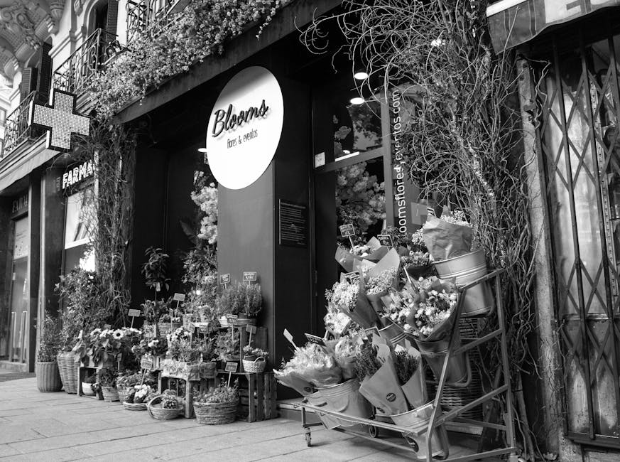 A florist shop named Bloom, showcasing a vibrant floral display outside its storefront on a city street. The arrangement features a variety of fresh flowers including roses, daisies, and lilies in dif