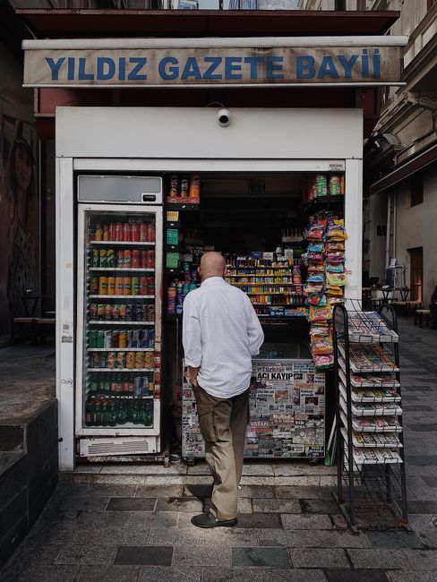 There is no image provided related to flowers, floral arrangements, or flower delivery content. The image shows a man in a white shirt and beige trousers standing outside a small kiosk or newsstand wi