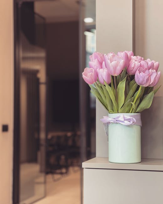 A bouquet of fresh pink tulips with closed petals and green leaves arranged in a pale green cylindrical vase wrapped with a light purple ribbon, placed on a white surface near a wall. The tulips are v
