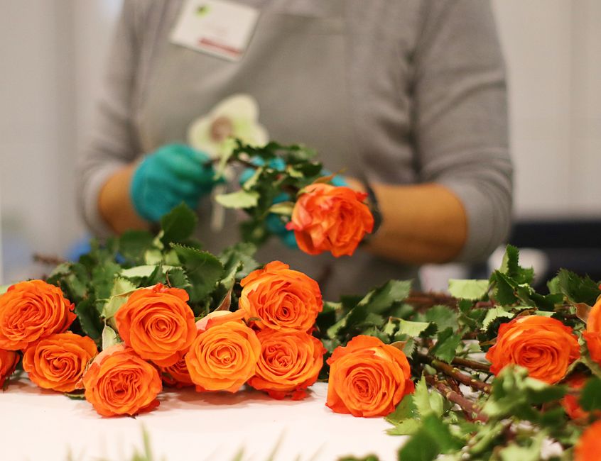 A florist, wearing a grey long-sleeved top and blue gloves, is arranging a floral bouquet with vibrant orange roses on a white work surface. The bouquet includes multiple fully bloomed roses with deli