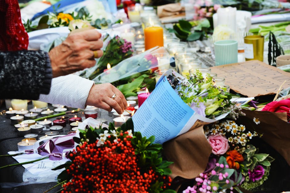 A close-up of a memorial floral tribute featuring a variety of fresh flowers, including pink roses, white daisies, and red berries, arranged in paper wrapping with purple, pink, and green hues. The bo