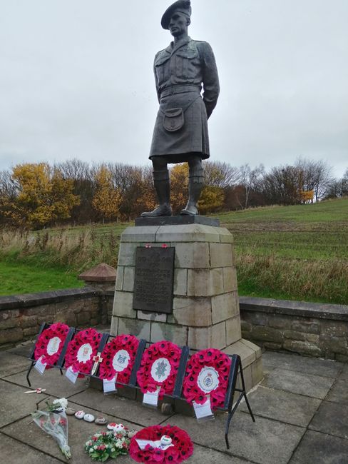 A bronze statue of a young girl in military uniform, standing atop a stone plinth with a commemorative plaque, is surrounded by five vibrant pink floral wreaths and smaller bouquets laid at its base, 