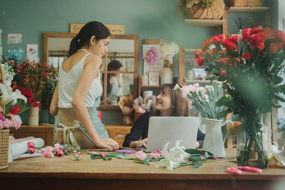 A woman florist in a white sleeveless top and beige apron engaging in a conversation with a female customer seated at a wooden table inside a floral shop. The customer is smiling and looking up at the