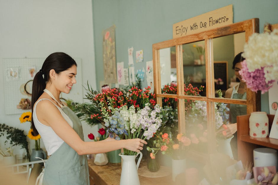 A young woman with dark hair tied back, wearing a white sleeveless top and a light green apron, is smiling as she arranges a bouquet of fresh flowers in a white ceramic vase at a flower shop. The bouq