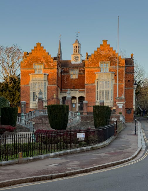 The image depicts a grand, historic red-brick building with a symmetrical façade featuring stepped gables, large bay windows, and a central clock tower with a weather vane on top, set against a clear