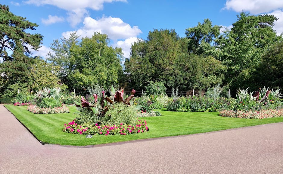 A well-maintained garden with vibrant flower beds featuring various flowering plants, including pink, purple, and white blooms, arranged around lush green lawns. In the foreground, a circular flower b