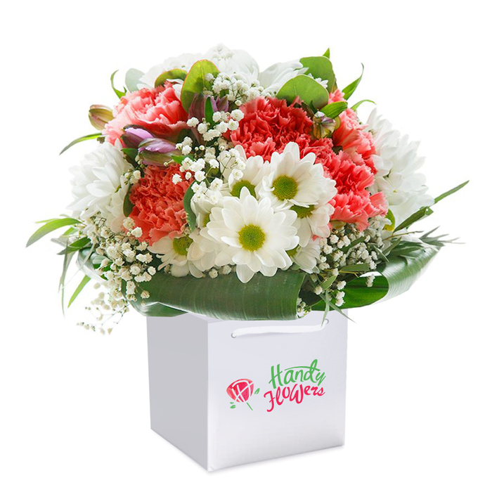 This meticulously crafted bouquet, standing proudly within a modern square white gift bag, embodies the fresh elegance and heartfelt artistry typical of Harrow on the Hill florists. The focal point is a lush interplay of coral, peach, and vivid red carnations, each bloom with intricately frilled petals lending a tactile richness and vibrant depth. Surrounding these are snowy white daisies, their bright yellow-green centers sparkling with cheerful energy, interspersed with airy sprigs of delicate baby's breath that seem to drift like gentle clouds through the composition. From the left, subtle hints of purple-tinged buds peek through-perhaps poised to flower, suggesting the quiet anticipation of spring mornings by Grove Hill. The arrangement is supported and defined by generous swathes of broad, glossy dark green foliage, wrapping the base in an elegant, natural collar that reflects the verdant charm of local parks and gardens. The crisp white gift bag, complete with sturdy ribbon handles, provides a contemporary finish and effortless delivery, perfect for gifting. Set against a flawless white background, every nuance of texture and shade is clear, inviting the eye to linger over the refined details. This radiant creation is suitable for marking a milestone, showing appreciation, or simply brightening a day-always a thoughtful choice for flower delivery throughout Harrow on the Hill.