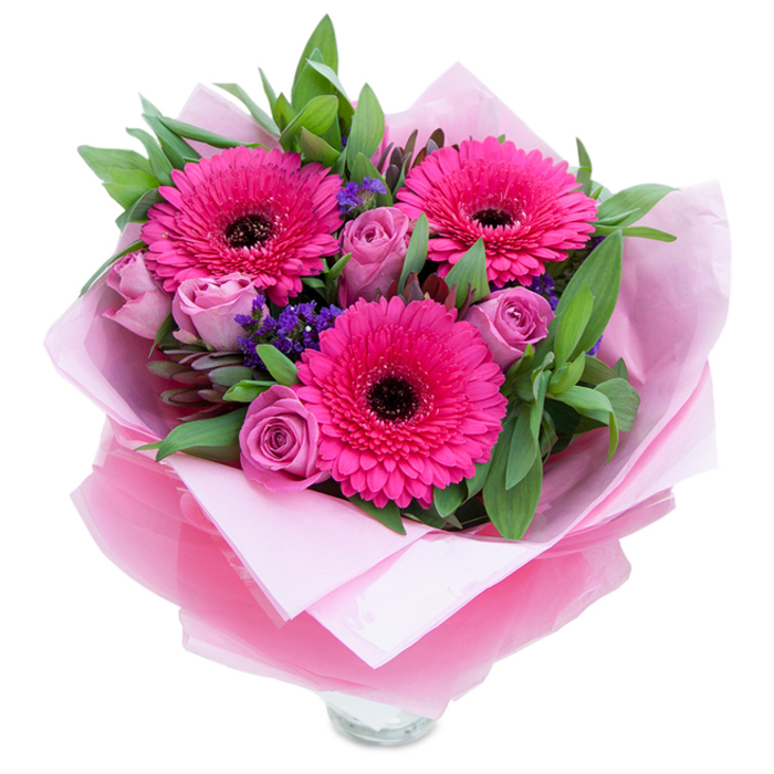 I picture this bouquet as a careful conversation between boldness and gentleness: three striking fuchsia Gerbera blooms form a confident trio, each with a dark central disk that anchors the eye and emphasizes the vibrancy of the petals. Nestled in their shadow are softer, dusty-rose to lavender-pink roses, still in bud and offering a velvety softness that calms the gerberas' drama. Tiny sprays of purple statice thread through the arrangement, their papery clusters providing a delicate, textural counterpoint and a cool violet note that deepens the colour story. Surrounding all of this, substantial green foliage-broad, glossy leaves that read like salal and the narrow, silvery-green hints of eucalyptus-creates a full, living backdrop and contributes a crisp, slightly resinous scent. The bouquet is wrapped with care: an inner layer of pale pink matte tissue cradles the stems while an outer wrap of translucent fuchsia-pink cellophane adds a gentle shimmer and holds the design together. You can see the top rim of a clear glass vase at the base, suggesting immediate display and the life-giving water beneath. The overall effect is intimate and uplifting, a piece I would send to mark a birthday, an anniversary, or simply to lift someone's spirits; I envision it brightening a bedside table in a cottage near Headstone Manor or greeting a neighbour returning from the shops on Pinner Road, a small, fragrant reminder of thoughtfulness and care.