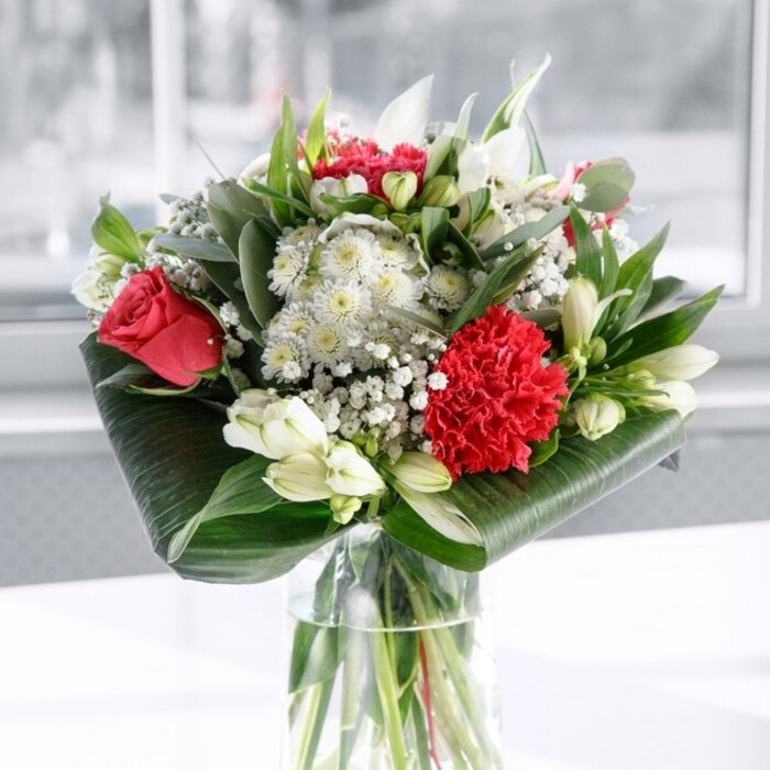In this image, a sophisticated red and white bouquet rises from a clear cylindrical glass vase, the stems sharply trimmed and fanned out below the water like a careful sketch of green lines. The arrangement is dominated by sumptuous deep red roses, their petals velvety and layered, each bloom poised somewhere between bud and full flower, suggesting romance that feels both timeless and freshly expressed. Nestled among them, a large crimson carnation unfolds in intricate ruffles, its densely packed petals adding a sense of rich volume and almost fabric-like texture. Around these passionate reds, a collection of white and cream flowers provides softness and balance: white alstroemeria with slim, slightly curved petals marked by fine speckles, neat clusters of tiny white gypsophila that float lightly between the larger blooms, and several ivory lily buds, still closed but tall and sculptural, hinting at days of beauty yet to come. The greenery has been handled with a trained florist's eye. Broad, glossy leaves are wrapped and curled at the base, forming a structured, modern collar that frames the flowers like a living pedestal, while slimmer, eucalyptus-style foliage and pointed green leaves weave through the bouquet, introducing tonal shifts from deep forest to silvery green. The arrangement is set on a white surface, against a softly diffused, bright background that recalls natural light in a Harrow on the Hill townhouse overlooking the leafy paths towards Harrow Recreation Ground. The overall impression is elegant yet welcoming, a centerpiece that feels as suitable for a refined anniversary dinner as for brightening a hallway or office reception, carrying with it the quiet message of appreciation, affection, and celebration of shared moments.